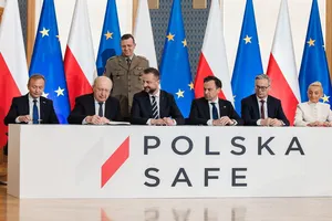 Grupa przedstawicieli władz państwowych siedzi przy białym stole z napisem „POLSKA SAFE”, za nimi stoją flagi Polski i Unii Europejskiej oraz oficer w mundurze.