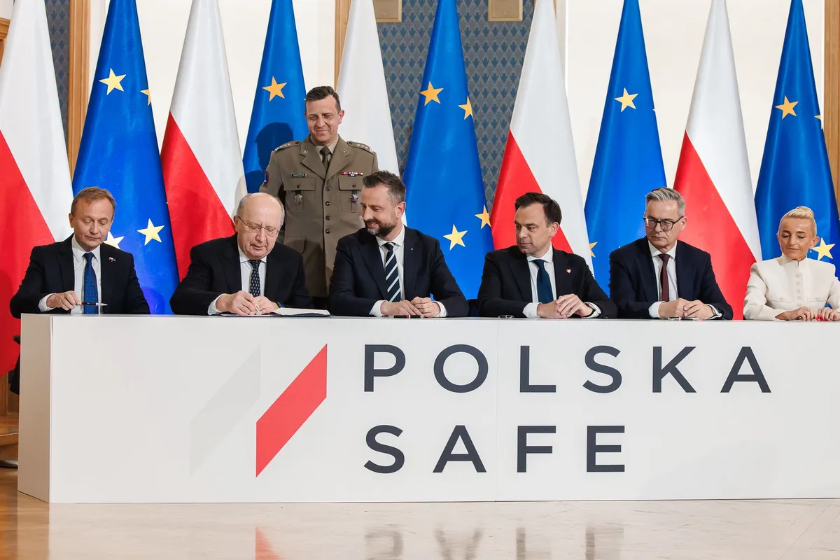 Grupa przedstawicieli władz państwowych siedzi przy białym stole z napisem „POLSKA SAFE”, za nimi stoją flagi Polski i Unii Europejskiej oraz oficer w mundurze.