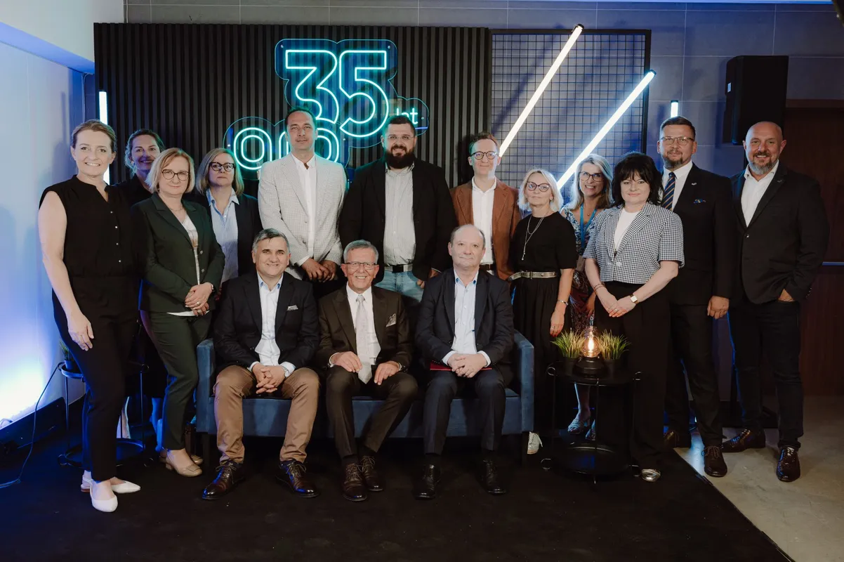 Grupa pracowników w formalnych strojach pozuje do zdjęcia w nowoczesnym wnętrzu; kilka osób siedzi na kanapie, pozostali stoją za nimi. W tle świeci neonowy napis „35 lat” oraz dekoracyjne lampy, podkreślające jubileuszowy charakter.
