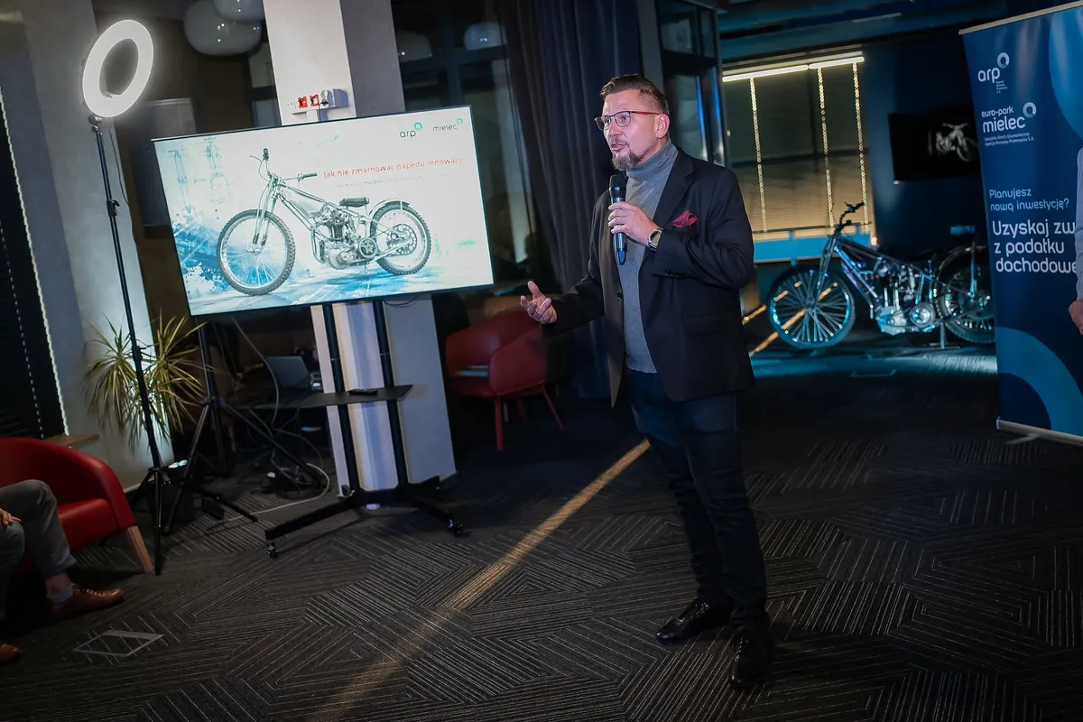 Prelegent występuje z mikrofonem podczas wydarzenia Benefis FIS, stojąc obok ekranu z prezentacją oraz roll-upu SSE EURO-PARK MIELEC. W tle widoczna jest ekspozycja motocykla, a przestrzeń wydarzenia ma kameralny, konferencyjny charakter