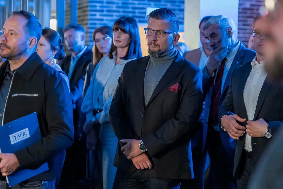 Uczestnicy wydarzenia Benefis FIS stoją w sali podczas części oficjalnej, słuchając programu spotkania w niebieskim oświetleniu