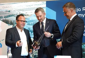 Trzech mężczyzn w garniturach ogląda z bliska szklaną statuetkę pamiątkową podczas konferencji.