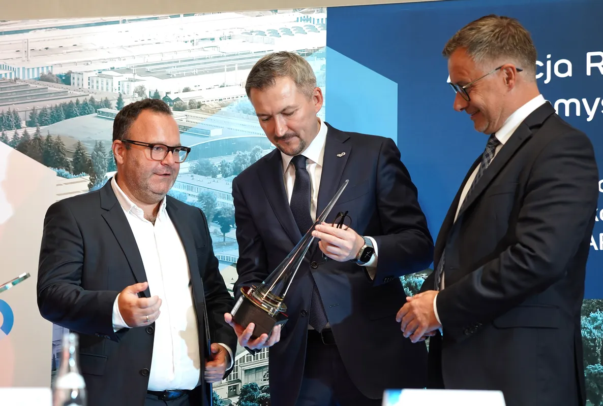 Trzech mężczyzn w garniturach ogląda z bliska szklaną statuetkę pamiątkową podczas konferencji.