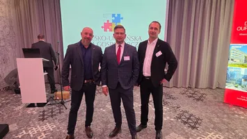 Trzech uczestników Krakowskiego Business Mixera pozuje na tle ekranu z logo wydarzenia oraz roll-upu promocyjnego. W tle widoczne są elementy sali konferencyjnej, w tym mównica, sprzęt techniczny oraz zasłony.