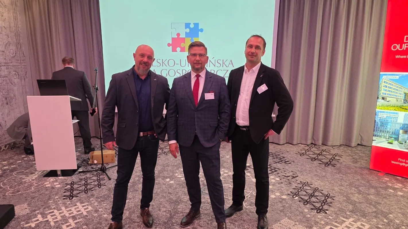 Trzech uczestników Krakowskiego Business Mixera pozuje na tle ekranu z logo wydarzenia oraz roll-upu promocyjnego. W tle widoczne są elementy sali konferencyjnej, w tym mównica, sprzęt techniczny oraz zasłony.