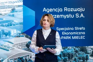 Magdalena Suraj stojąca z tabletem w dłoniach na tle ścianki z logo Agencji Rozwoju Przemysłu S.A. i Specjalnej Strefy Ekonomicznej EURO-PARK MIELEC.