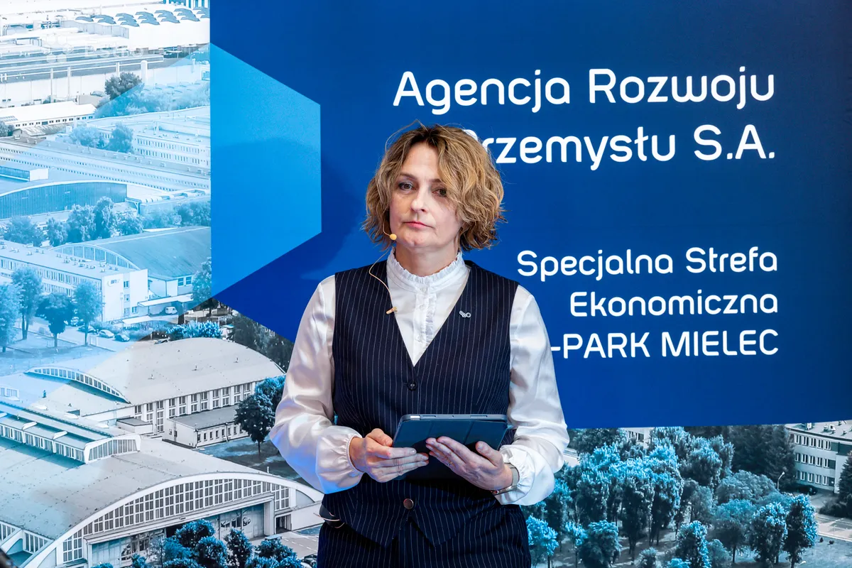 Magdalena Suraj stojąca z tabletem w dłoniach na tle ścianki z logo Agencji Rozwoju Przemysłu S.A. i Specjalnej Strefy Ekonomicznej EURO-PARK MIELEC.