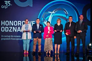 Siedem osób stoi na scenie podczas uroczystości wręczenia odznaczeń, trzymając czerwone teczki i medale. W tle widoczny napis „35 lat arp” oraz grafika ekranu w niebieskiej tonacji.