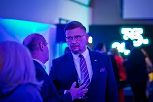 Mężczyzna w okularach i eleganckim garniturze rozmawia z uczestnikami eventu. W tle widoczne są inne osoby oraz rozmyty, świecący neon jubileuszowy „35 lat”.