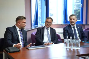 Trzech mężczyzn w garniturach rozmawiających przy stole konferencyjnym, mężczyzna po lewej gestykuluje podczas wypowiedzi.