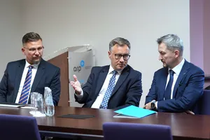 Mężczyzna w okularach gestykuluje dłonią podczas rozmowy z dwoma innymi mężczyznami przy stole konferencyjnym.