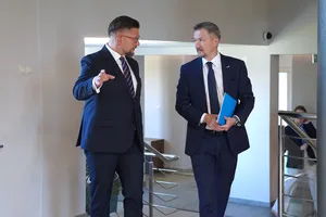 Dwaj mężczyźni w garniturach idący i rozmawiający ze sobą na korytarzu budynku.