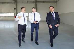 Alt: Trzech mężczyzn idących i rozmawiających wewnątrz pustej, przestronnej hali przemysłowej.