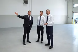 Mężczyzna w ciemnym garniturze i okularach gestykuluje dłonią, wskazując wnętrze pustej hali dwóm towarzyszącym mu mężczyznom.
