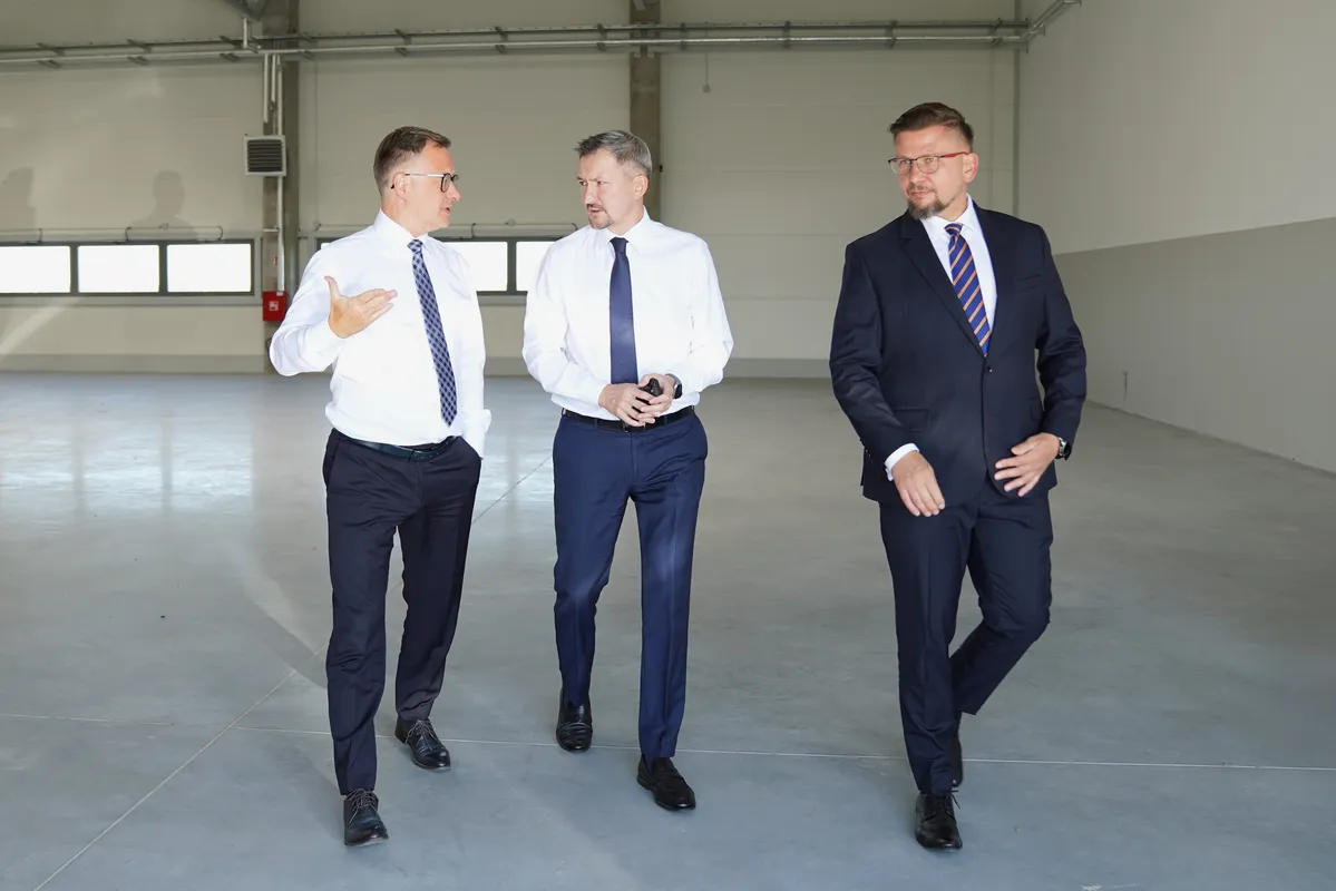 Alt: Trzech mężczyzn idących i rozmawiających wewnątrz pustej, przestronnej hali przemysłowej.