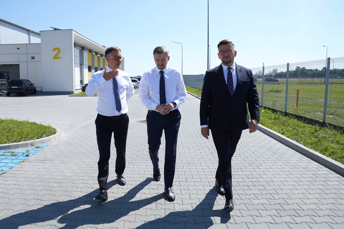 Trzech mężczyzn w formalnych strojach idących wybrukowaną alejką na terenie strefy przemysłowej.