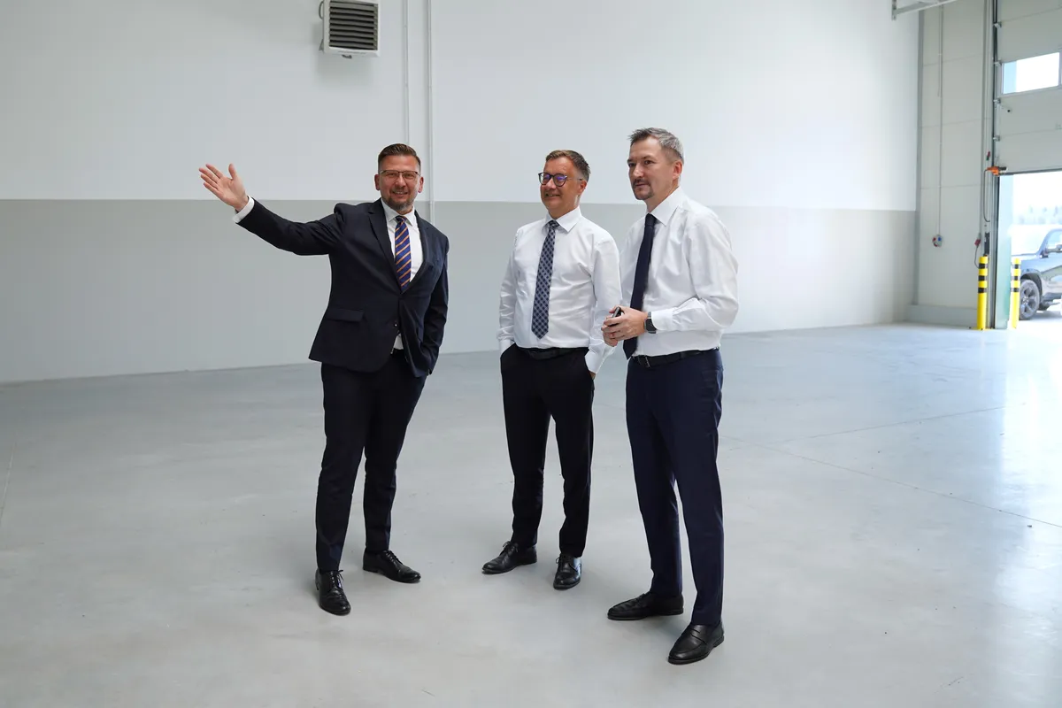 Mężczyzna w ciemnym garniturze i okularach gestykuluje dłonią, wskazując wnętrze pustej hali dwóm towarzyszącym mu mężczyznom.