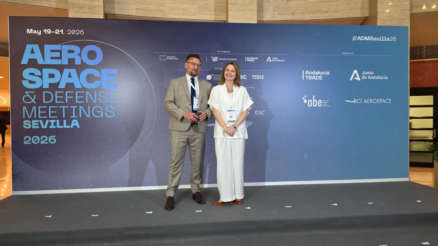 Mężczyzna i kobieta stoją na scenie przed ścianką z logotypami organizatorów i hasłem Aerospace & Defense Meetings Sevilla 2026.