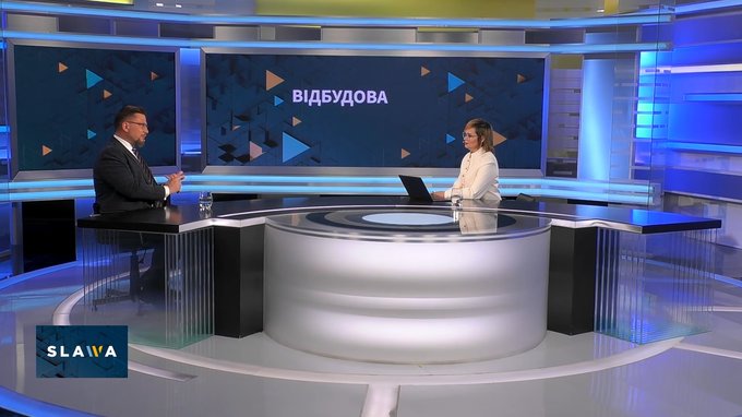 Nowoczesne studio telewizyjne podczas programu publicystycznego. Po lewej stronie przy biurku siedzi mężczyzna w garniturze, po prawej dziennikarka w jasnym żakiecie pracująca przy laptopie. W tle duży ekran z napisem „ВІДБУДОВА” oraz niebiesko-pomarańczo